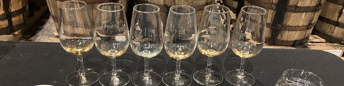 Six stemmed tasting glasses lined up in front of wooden whiskey casks.